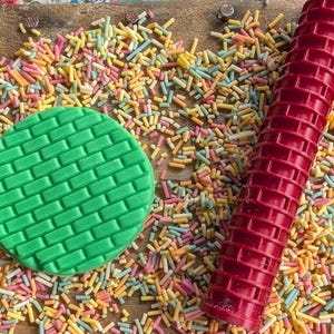 May include: A green, circular fondant stamp with a brick pattern is next to a red textured rolling pin. Colourful sprinkles are scattered across a wooden surface. The scene is vibrant and playful.