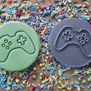 May include: Two round cookie cutters, shaped like game controllers, one green and one grey, on a wooden surface with colourful sprinkles. The controllers feature a cross and button details.