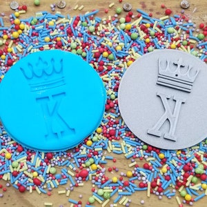 May include: Two round cookie cutters, one blue and one gray, each featuring a crown and the letter "K". The cutters are surrounded by colorful sprinkles, suggesting a baking theme.