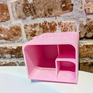 May include: A light pink desk organizer with a rectangular shape and a small shelf inside. The top of the organizer has the word "SPACE" embossed on it. The organizer is made of a smooth, solid material.