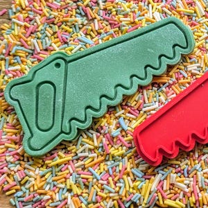May include: A green saw-shaped cookie cutter and a red saw-shaped cookie cutter are displayed on a bed of colorful sprinkles. The green cutter has a handle and a blade with jagged teeth. The red cutter is a partial view.