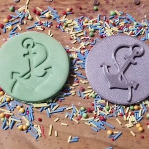 May include: Two round, embossed objects with anchor designs. One is light green, the other is silver. Colorful sprinkles surround the objects. Nautical-themed craft supplies or decorations.