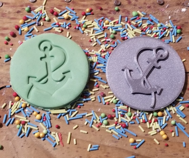 May include: Two round, embossed objects with anchor designs. One is light green, the other is silver. Colorful sprinkles surround the objects. Nautical-themed craft supplies or decorations.