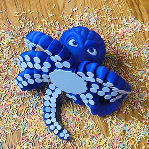 May include: A blue octopus figurine with white suckers on its tentacles. The octopus has cartoon-like eyes and a light blue belly. It is surrounded by colorful sprinkles on a wooden surface.