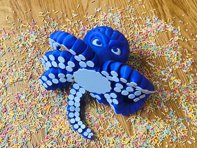 May include: A blue octopus figurine with white suckers on its tentacles. The octopus has cartoon-like eyes and a light blue belly. It is surrounded by colorful sprinkles on a wooden surface.