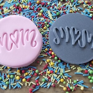 May include: Two round cookie stamps, one pink and one gray, with the word "MOM" in a cursive font with a heart in the center. The stamps are surrounded by colorful sprinkles on a wooden surface.