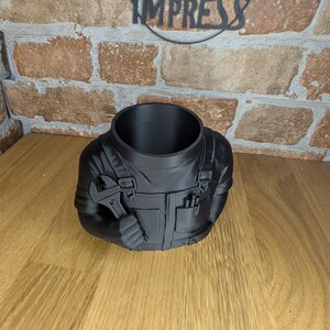 May include: Black planter shaped like a mechanic's torso, holding a wrench. The planter has a large opening at the top and features details like pockets and overalls. The background includes a brick wall and the word "IMPRESS".