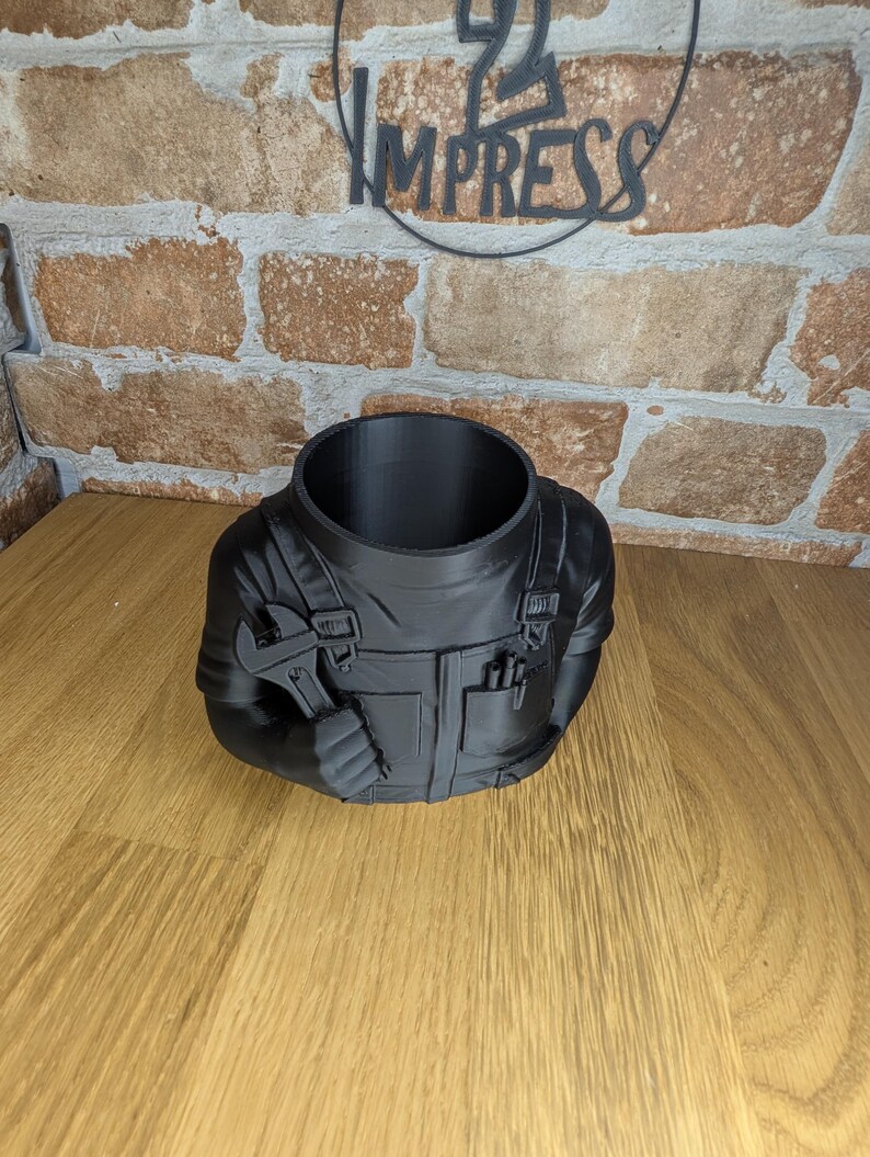 May include: Black planter shaped like a mechanic's torso, holding a wrench. The planter has a large opening at the top and features details like pockets and overalls. The background includes a brick wall and the word "IMPRESS".