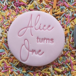 Puede incluir: Decoraci&oacute;n circular rosa claro para pastel con la frase "Alice turns One" en cursiva. Rodeada de coloridas chispas, ideal para celebrar un primer cumplea&ntilde;os.