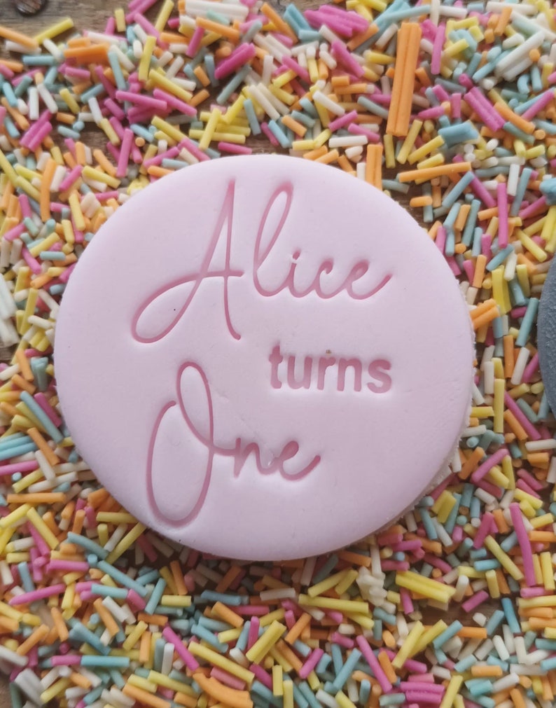 Puede incluir: Decoraci&oacute;n circular rosa claro para pastel con la frase "Alice turns One" en cursiva. Rodeada de coloridas chispas, ideal para celebrar un primer cumplea&ntilde;os.
