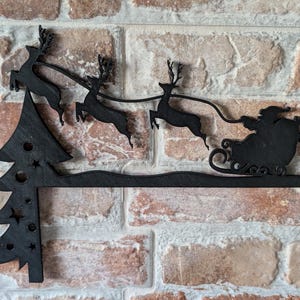 May include: Black wooden Christmas decoration featuring Santa Claus in a sleigh pulled by reindeer, with a Christmas tree on the left. The decoration is mounted on a brick wall, creating a festive holiday scene.