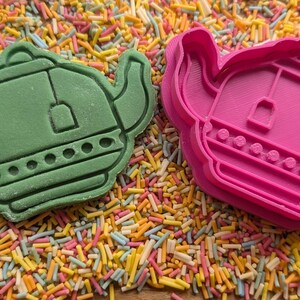 May include: A green biscuit and a pink biscuit cutter, both shaped like a teapot, sit on a bed of colourful sprinkles. The green biscuit is a finished product, while the pink one is the cutter.