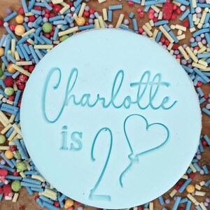 May include: A light blue round cookie with the text "Charlotte is 2" and a heart-shaped balloon. The cookie is surrounded by colorful sprinkles.