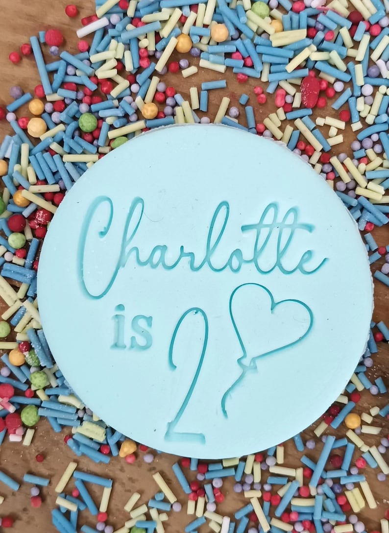 May include: A light blue round cookie with the text "Charlotte is 2" and a heart-shaped balloon. The cookie is surrounded by colorful sprinkles.