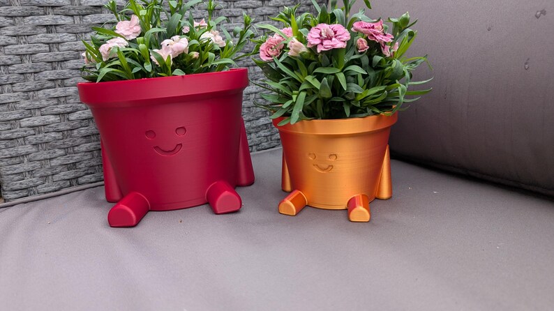 May include: Two whimsical, smiling flower pots, one red and one orange, each with attached legs. The pots are filled with green foliage and pink flowers. A fun and unique addition to any home or garden.