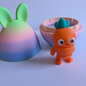 May include: A pastel rainbow colored Easter egg with bunny ears opens to reveal a small orange carrot character with green leaves and a smiling face.
