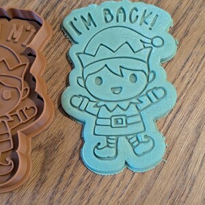 May include: A teal-colored cookie cutter in the shape of an elf with the text "I'M BACK!" above the elf's head. The elf is wearing a hat, belt, and shoes. A brown cookie cutter is visible on the left.