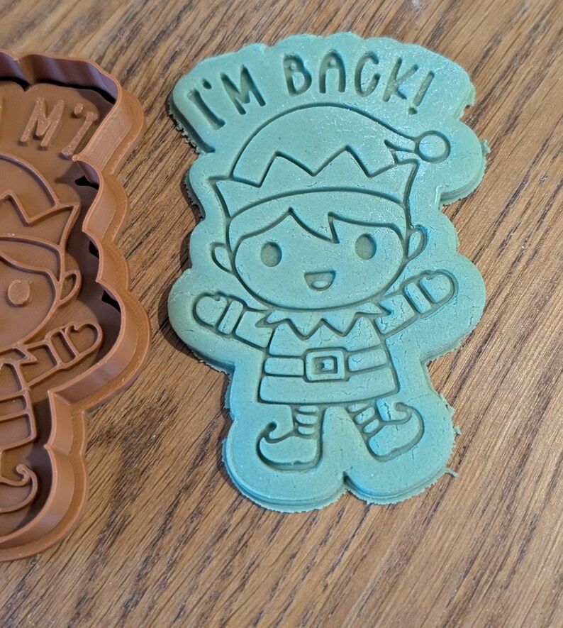 May include: A teal-colored cookie cutter in the shape of an elf with the text "I'M BACK!" above the elf's head. The elf is wearing a hat, belt, and shoes. A brown cookie cutter is visible on the left.
