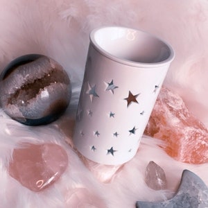 May include: White ceramic wax warmer with a star cut-out design. The warmer is surrounded by various pink and blue crystals.