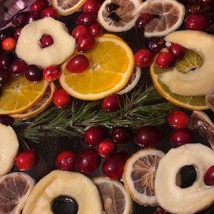 May include: A glass bowl filled with a red liquid, cranberries, orange and lemon slices, apple rings, and rosemary sprigs.