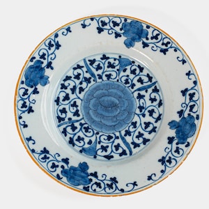 Puede incluir: Un plato de cerámica blanca con un diseño floral azul. El plato presenta un motivo floral central y un borde de flores y vides estilizadas. El borde tiene un ribete de color dorado.