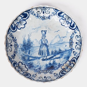 Può includere: Un piatto rotondo in ceramica blu e bianca antica con bordo smerlato. Il piatto presenta una scena di paesaggio dipinta a mano di una donna che trasporta una brocca, con alberi, montagne e dettagli floreali. Il piatto è un oggetto decorativo.