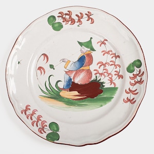 Pode incluir: Um prato de cerâmica branco com um desenho pintado em vermelho, verde e azul de uma pessoa com um chapéu verde sentada em uma rocha com flores ao redor.