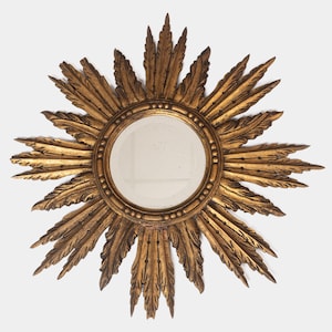 May include: A gold-colored sunburst mirror with a round, clear mirror center. The sunburst design features ornate, leaf-like rays radiating outwards. The frame has a textured, antique finish, adding a vintage aesthetic to the decorative wall mirror.