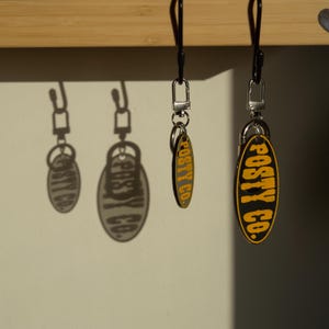 Puede incluir: Dos llaveros con etiquetas ovaladas negras y texto amarillo que dice "POSTY CO." cuelgan de cordones negros. Las etiquetas son negras con texto amarillo. Los llaveros cuelgan frente a una pared blanca con sombras.
