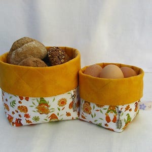 Könnte beinhalten: Zwei Stoffkörbe mit gelben, gesteppten Oberteilen und gemusterten Böden. Ein Korb enthält Brötchen, der andere Eier. Der gemusterte Stoff zeigt Zwerge, Blumen und Blätter in Orange, Grün und Weiß.