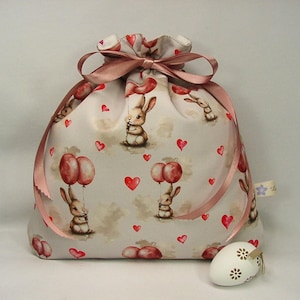 Könnte beinhalten: Ein Geschenkbeutel mit Kordelzug und einem sich wiederholenden Muster aus Hasen, die rote Luftballons und rote Herzen auf hellem Hintergrund halten. Der Beutel ist mit einem rosa Band gebunden. Ein Osterei mit floralen Designs liegt vor dem Beutel.