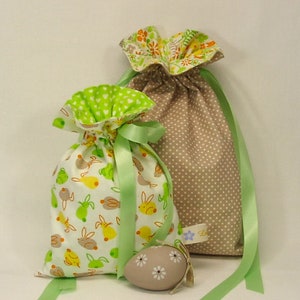 Könnte beinhalten: Zwei Stoffgeschenktüten mit grünen Schleifenbändern. Eine Tasche ist braun mit weißen Punkten und die andere ist weiß mit grünen und gelben Hasen und grünen Äpfeln. Ein braunes und weißes Osterei befindet sich im Vordergrund.