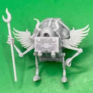 Könnte beinhalten: Hellgraue, 3D-gedruckte Fantasy-Figur mit einem kastenförmigen Körper, einem einzelnen Auge und einem lächelnden Mund. Sie hat kleine Flügel, Hörner und einen Stab mit gebogenem Ende. Die Figur steht auf zwei Beinen und hat bewegliche Arme.