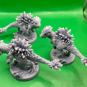 Peut inclure: Trois figurines de dinosaures grises avec des protubérances épineuses le long du dos et de la queue. Les dinosaures sont sur des socles ronds et se détachent sur un fond vert. Les figurines ont des textures détaillées.