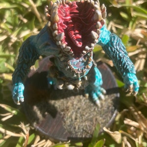 May include: A teal blue and white painted miniature of a monster with a large open mouth and sharp teeth. The monster is standing on a brown base.
