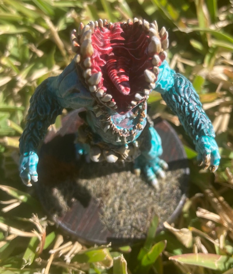 May include: A teal blue and white painted miniature of a monster with a large open mouth and sharp teeth. The monster is standing on a brown base.
