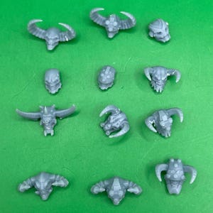 May include: A collection of gray, horned skull miniatures, each with unique horn and facial designs. The skulls are arranged on a bright green surface, showcasing various shapes and sizes.