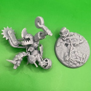 Könnte beinhalten: Eine graue, detaillierte Miniaturfigur einer mechanischen Kreatur mit einem sägeartigen Arm und einer kreisförmigen Waffe. Die Figur ist dynamisch dargestellt, mit einem separaten, runden Sockel mit einer Kettenstruktur.