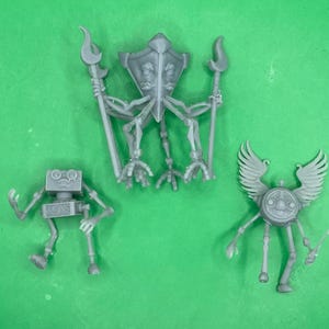 Könnte beinhalten: Drei graue Roboterfiguren aus Kunststoff sind auf einer grünen Oberfläche angeordnet. Eine Figur hat einen schildartigen Kopf und hält zwei Speere. Eine andere ist ein kastenförmiger Roboter mit quadratischem Kopf. Die dritte hat Flügel und einen kreisförmigen Körper.