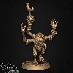 May include: A bronze-coloured miniature figure of a monster with multiple arms, a wide, toothy grin, and a long tongue. The figure is holding a staff-like object with a similar monster head on top. The base of the figure has the text "BATTLE YAK MINIATURES".