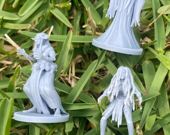 May include: Three light gray miniature fantasy figures are arranged on green grass. The figures depict characters in flowing robes and dresses, with one figure raising its arms. The figures are on circular bases.