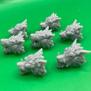 May include: Seven gray wolf head figurines with detailed fur and open mouths are arranged on a green surface. The wolf heads are facing different directions, showcasing their intricate design and texture. These miniature sculptures are likely for crafting or decorative purposes.