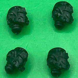 May include: Four small, black, helmet-shaped objects are arranged against a bright green background. The helmets have detailed features, including visors and other structural elements. The objects appear to be miniature replicas or components.