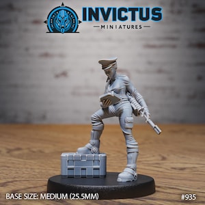 May include: A gray miniature figure of a person in a military uniform, holding a tablet and a rifle, standing on a crate. The figure is on a black base. The text "INVICTUS - MINIATURES" is at the top. Base size: Medium (25.5mm).