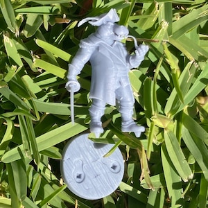 May include: A gray 3D printed miniature figurine of a pirate wearing a hat and holding a pipe. The figurine is standing on a round base.