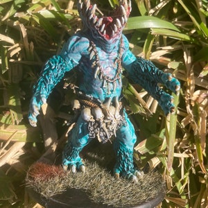 May include: A blue and white painted miniature figure of a creature with a large mouth and sharp teeth. The figure is wearing a bone necklace and a bone belt. The figure is standing on a base with brown and green grass.