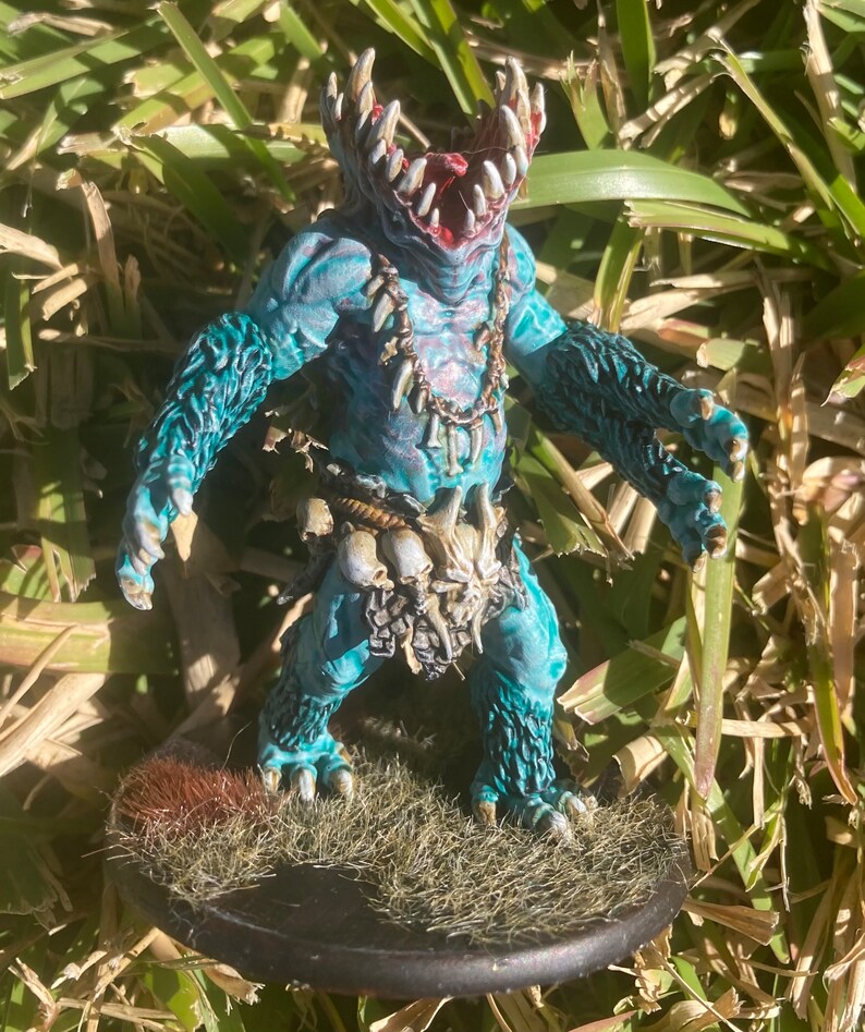 May include: A blue and white painted miniature figure of a creature with a large mouth and sharp teeth. The figure is wearing a bone necklace and a bone belt. The figure is standing on a base with brown and green grass.