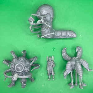 Könnte beinhalten: Eine Sammlung grauer, unbemalter Miniaturfiguren. Das Set beinhaltet ein wurmartiges Wesen mit skelettartigen Armen, eine stachelige Kugel mit einem Schädel, eine humanoide Figur und ein Wesen mit gebogenen Hörnern und mehreren Beinen. Die Figuren befinden sich auf einer grünen Oberfläche.