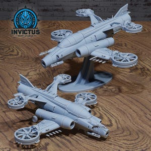 May include: Two gray miniature aircraft models with multiple propellers and detailed designs. One model is elevated on a stand. The Invictus Miniatures logo is visible in the upper left corner. The models are on a wooden surface.