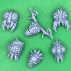 May include: A collection of six unpainted grey resin miniature heads and appendages featuring alien-like designs with multiple eyes, textured surfaces, sharp mandibles, and antler-like protrusions, arranged in a scattered pattern.
